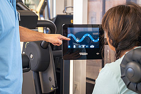 Mann zeigt auf Touchscreen eines Fitnessgeräts vor Frau im Fitnessstudio
