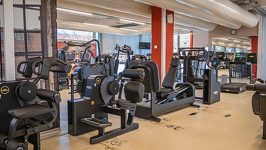 NEU! Premium-Training mit Technogym Innenansicht eines Fitnessraums mit mehreren Kraft- und Cardiogeräten, großen Fenstern und hellen Deckenleuchten.
